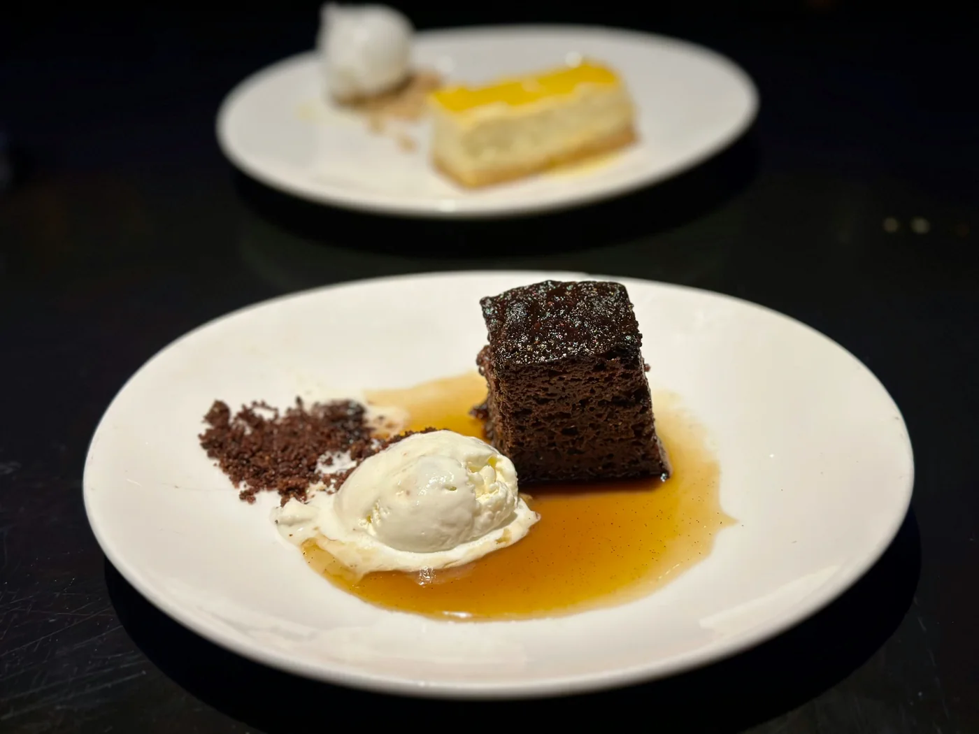 A square of sticky toffee pudding on a dark plate with toffee sauce and a scoop of ice cream