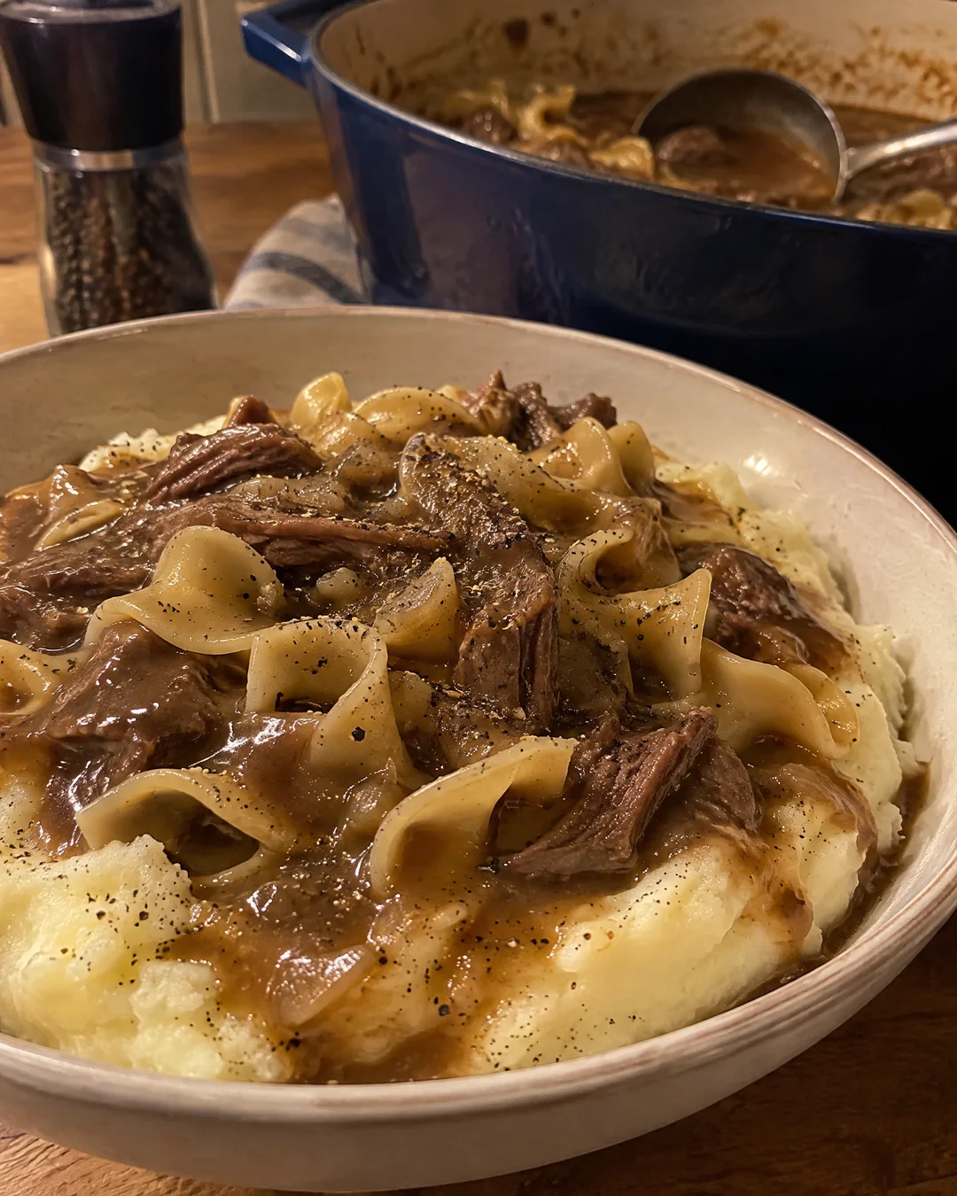 A bowl of mashed potatoes with a dent filled with beef and noodles, the pot and ladle just behind