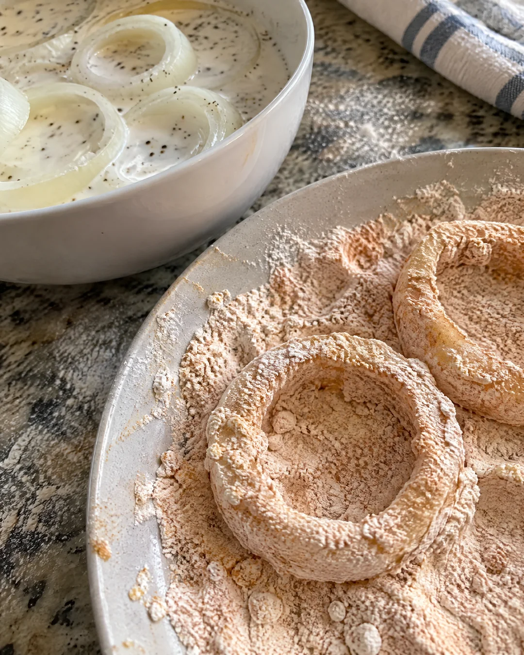 Thick onion rings soaking in buttermilk next to a dish of seasoned flour