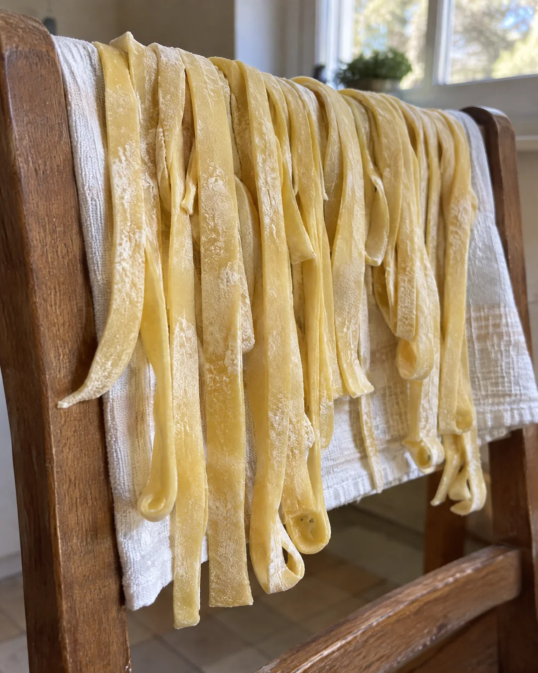 Fresh cut egg noodles drying on a flour-dusted dish towel hung over the back of a wooden kitchen chair