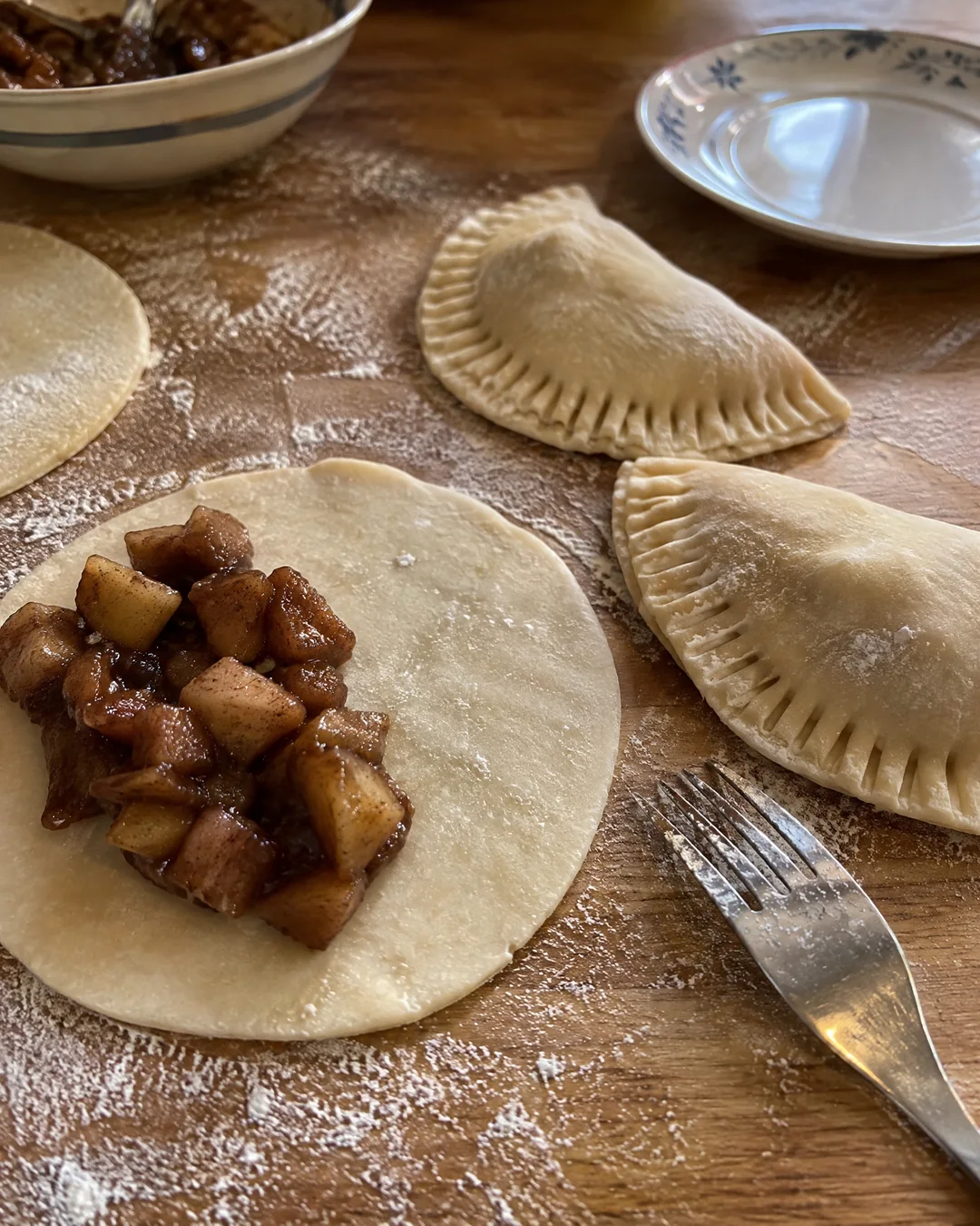 Rolled dough circles on a floured board with dried apple filling on one half and fork-crimped half moons beside them