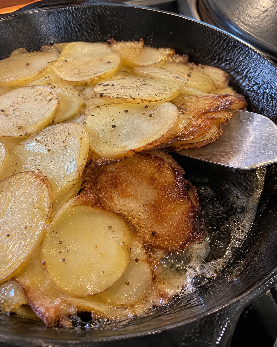 Sliced potatoes frying uncovered in a cast iron skillet, the lid just removed and the bottom layer turning deep brown