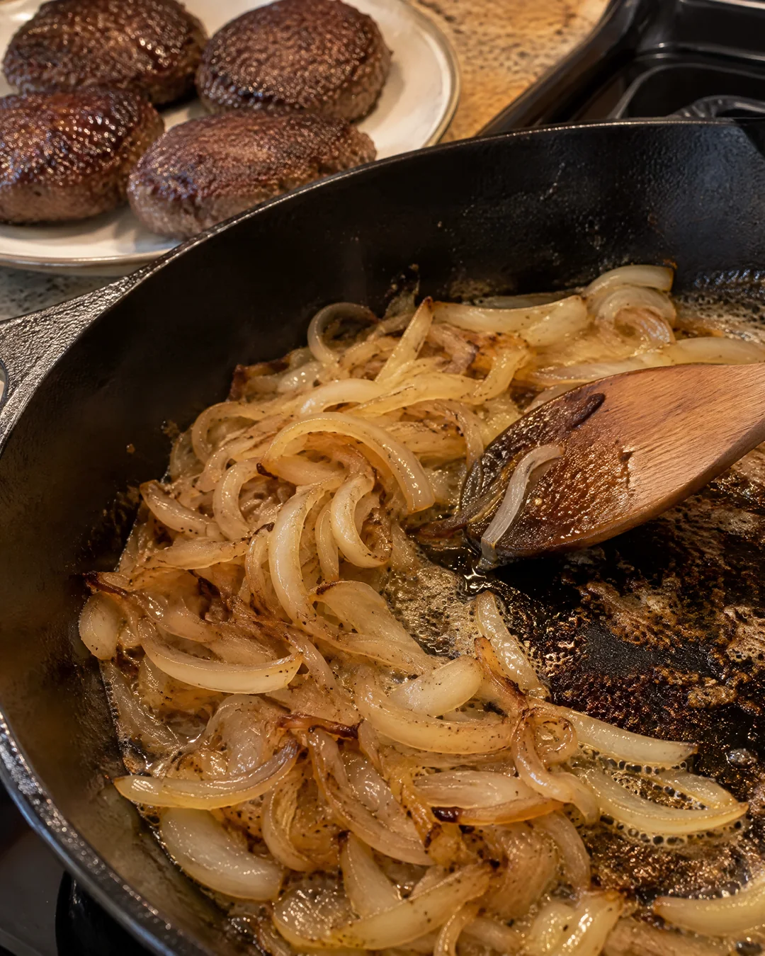 Sliced onions cooking slowly in a cast iron skillet, lifting the brown bits left by the hamburger patties