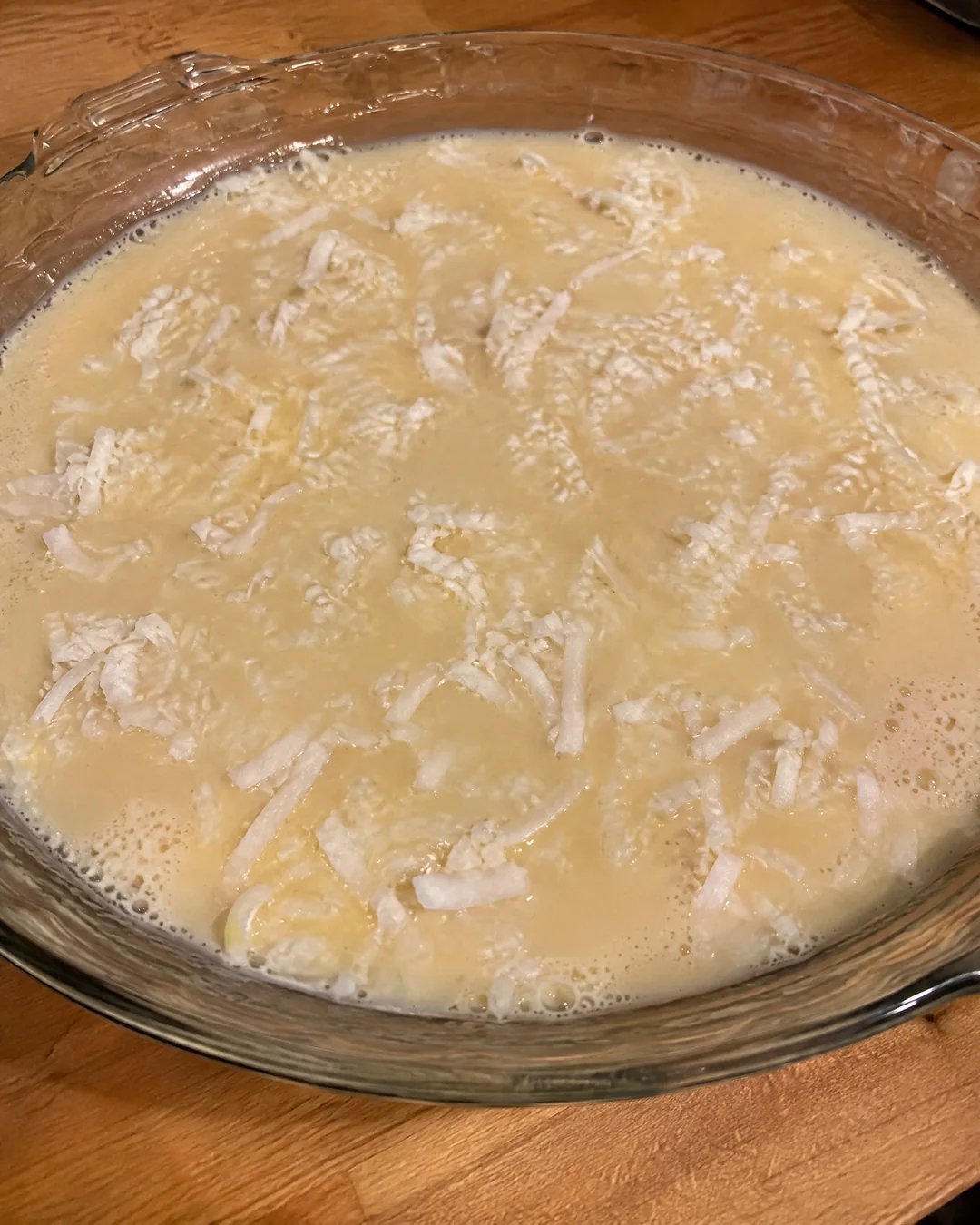 Thin coconut pie batter poured into a buttered pie plate with no crust, coconut flakes floating on top