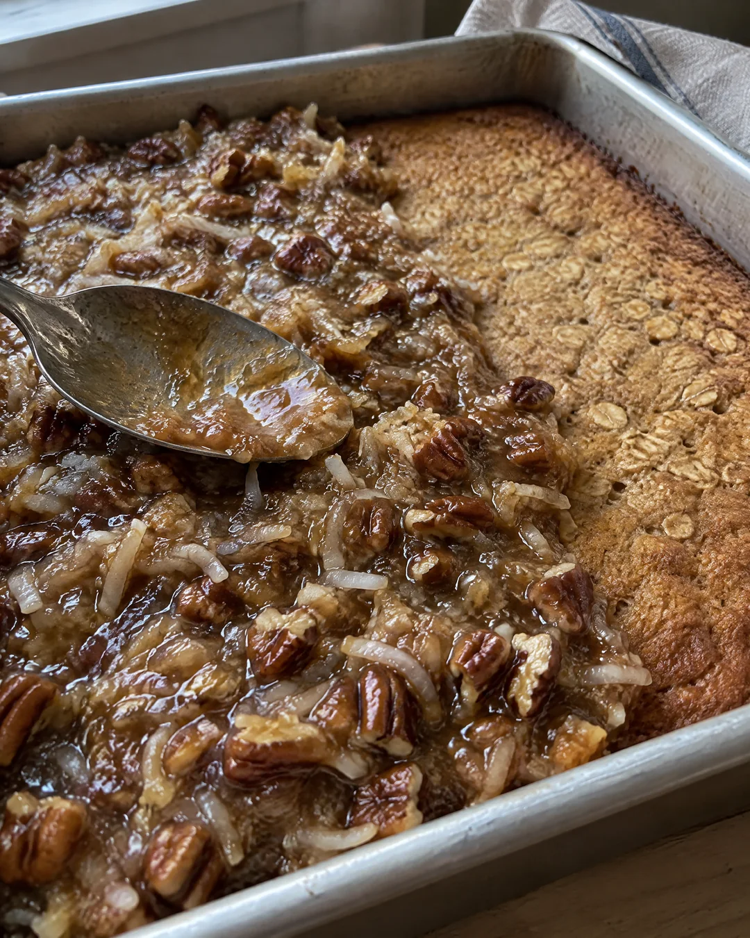 Coconut and pecan brown sugar topping being spread with the back of a spoon over a hot oatmeal cake in a metal pan before broiling