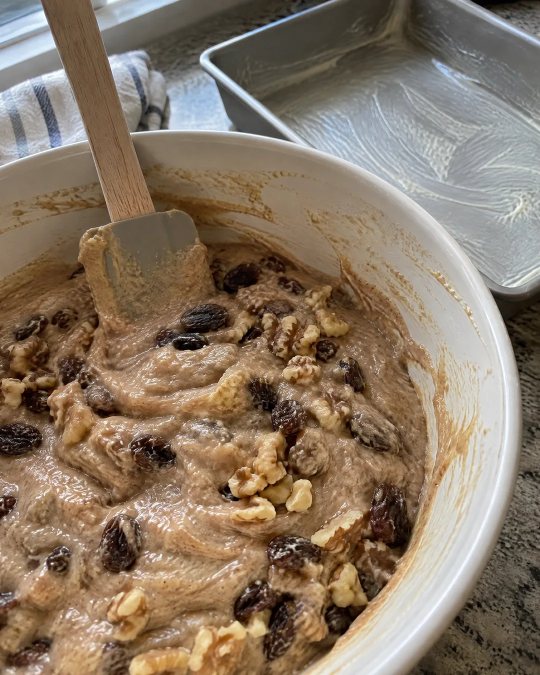 Thick spiced applesauce cake batter with raisins and walnuts being folded together in a mixing bowl