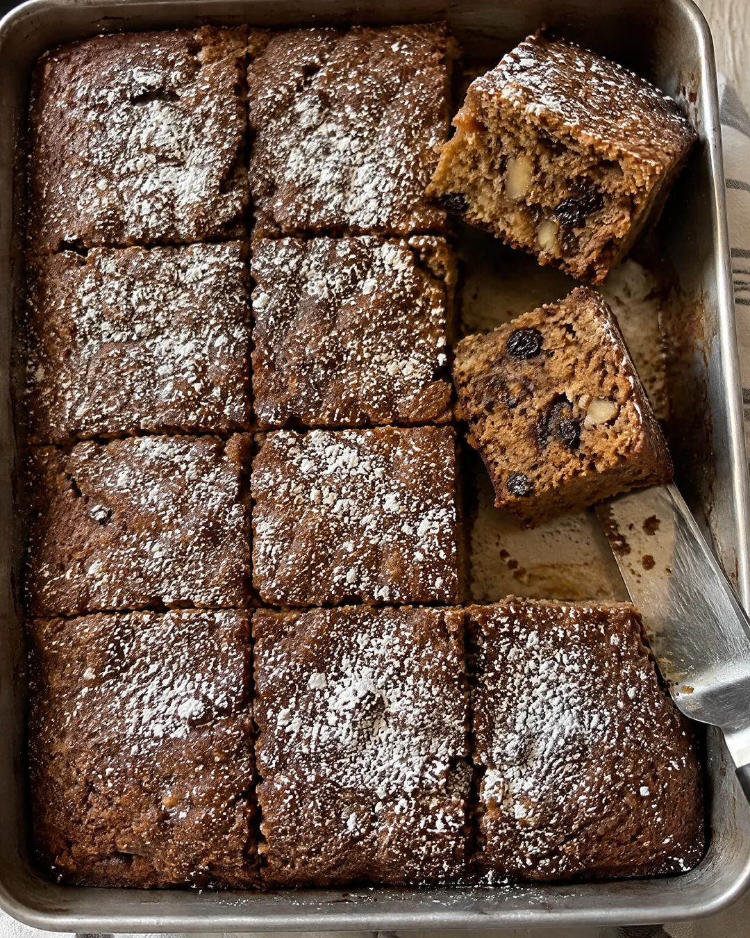 A 9 by 13 pan of dark applesauce cake dusted with powdered sugar and cut into squares, seen from above with two squares lifted out