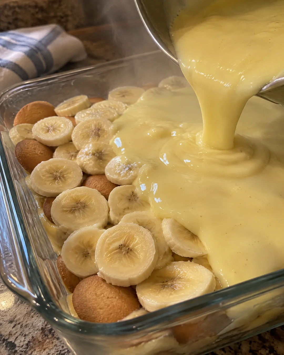 Hot cooked custard being poured over layers of vanilla wafers and banana slices in a glass dish