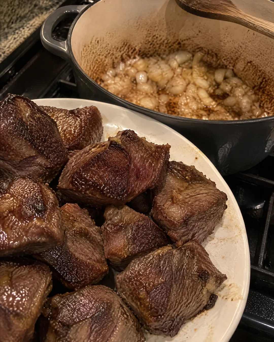 Flour-coated chunks of beef chuck browned dark on all sides piled on a plate beside the heavy pot where onions cook in the browned bits