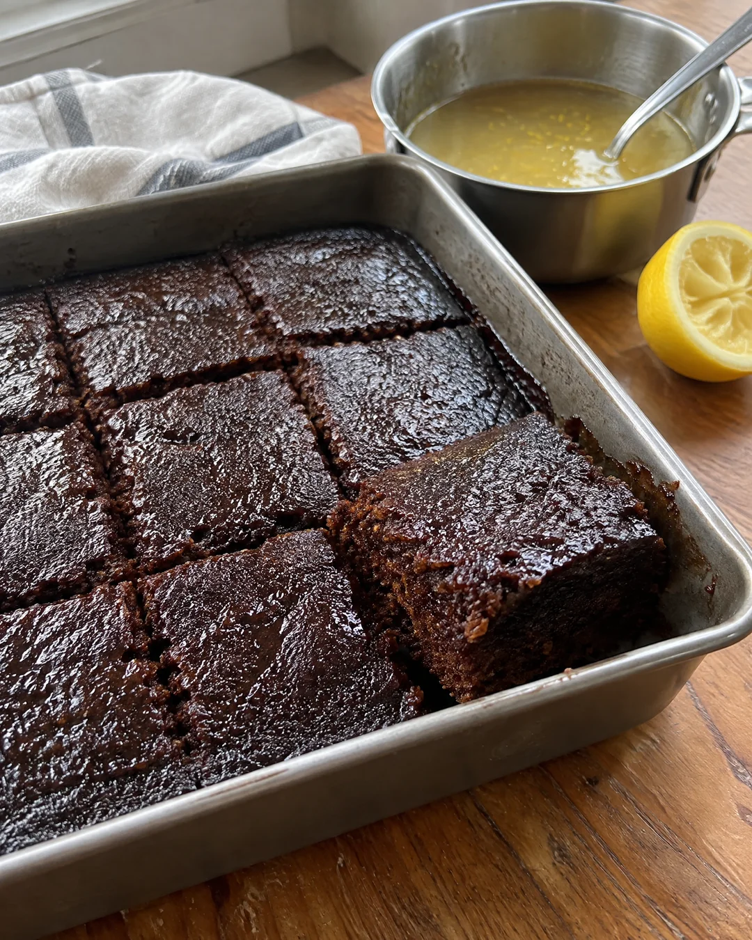 A square pan of dark old-fashioned gingerbread cut into nine squares with a small saucepan of warm lemon sauce beside it
