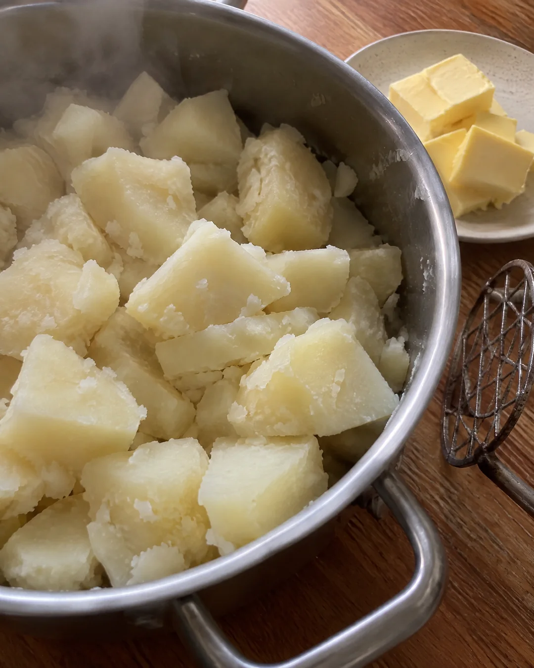Boiled russet potatoes drying in the hot pot after draining, floury at the edges, with a wire masher and butter waiting