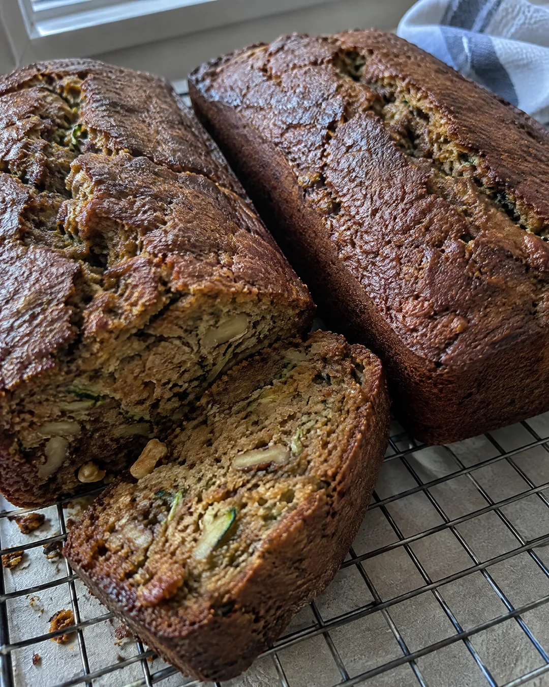 Two dark loaves of zucchini bread cooling on a wire rack, one with the end cut off to show the moist green-flecked crumb
