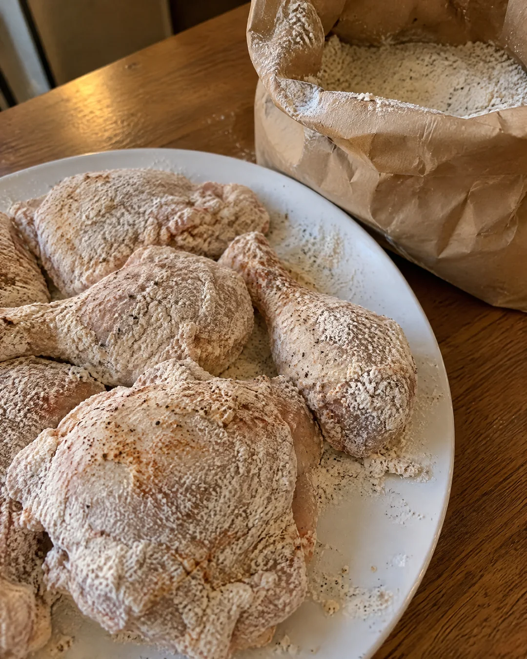 Flour-coated chicken pieces resting on a plate beside the open brown paper grocery bag of seasoned flour