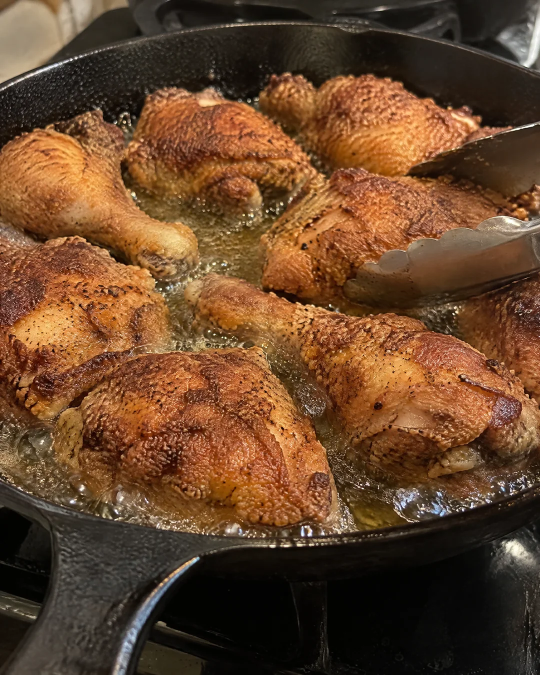 Fried chicken pieces frying uncovered in a black cast iron skillet, crusts deep golden brown, tongs turning a thigh