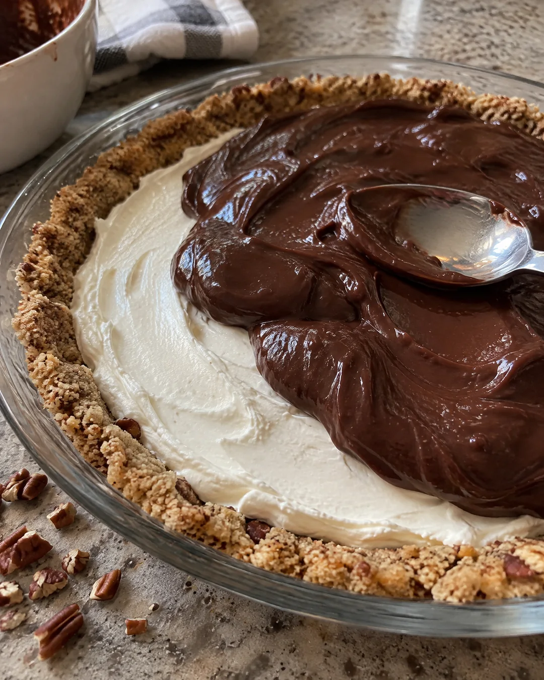 Chocolate pudding being spread over the white cream cheese layer in a pecan shortbread crust