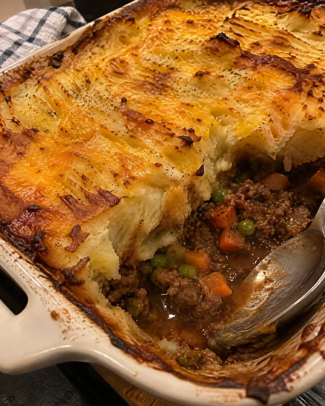A baked shepherd's pie in its dish with one corner scooped out, showing beef, peas and carrots in gravy under the browned potato