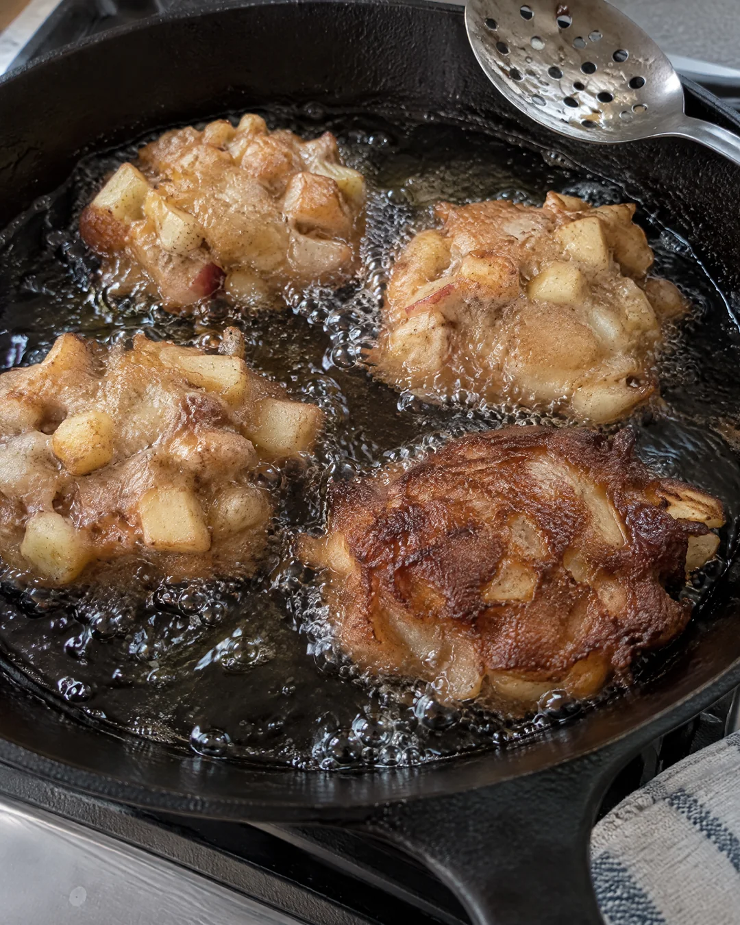 Spoonfuls of thick apple fritter batter frying in an inch of hot oil in a cast iron skillet, edges bubbling golden