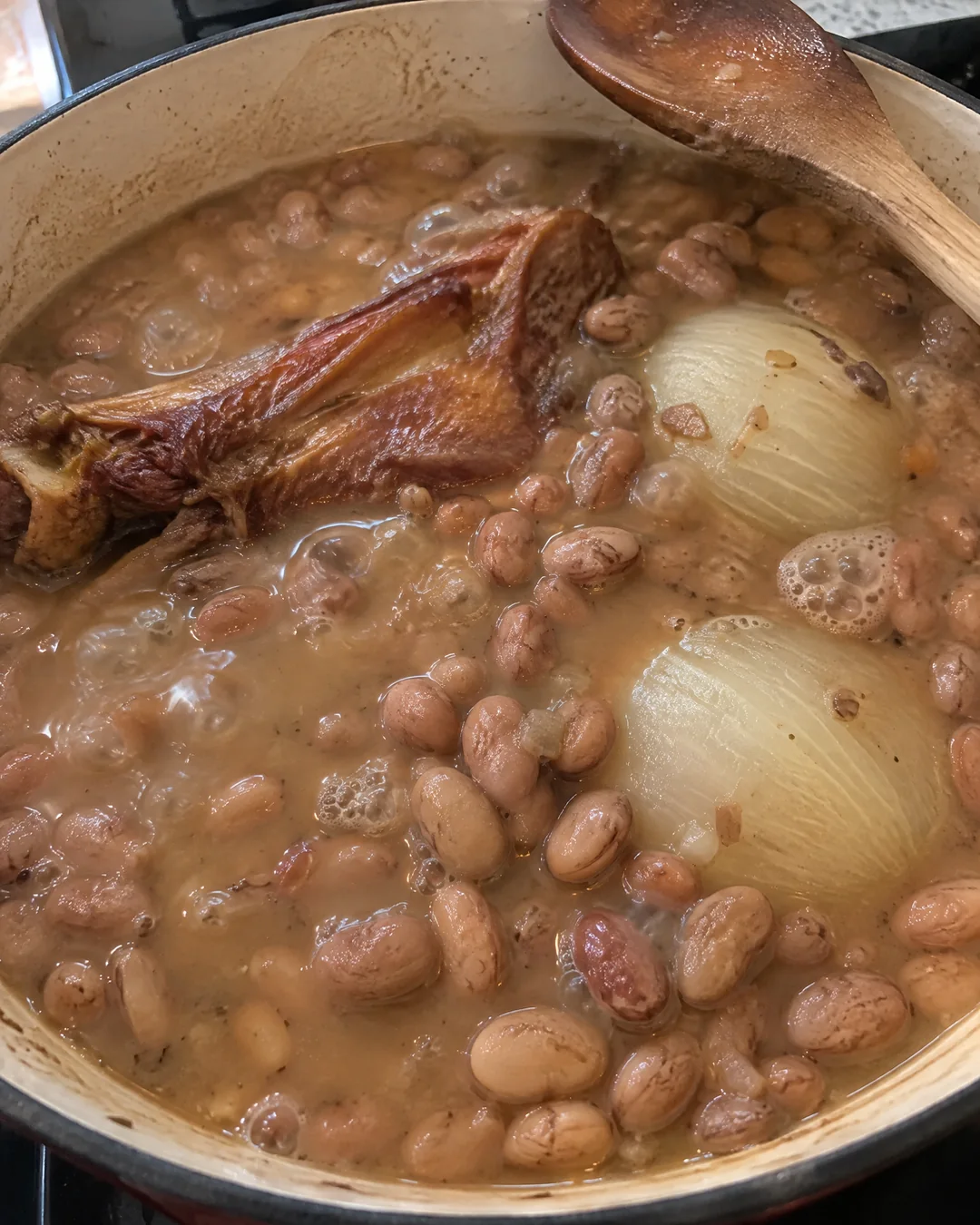 A heavy pot of pinto beans at a slow simmer with a smoked turkey wing and onion halves in a cloudy broth