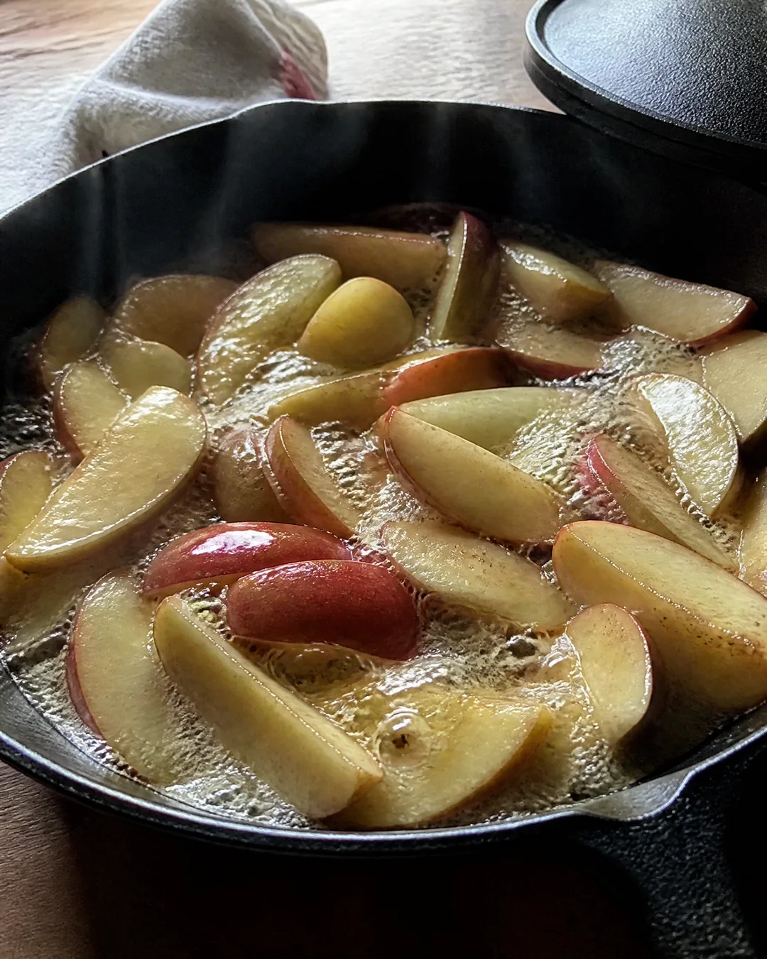 Thick apple wedges with red skins steaming in butter in a cast iron skillet before the sugar goes in