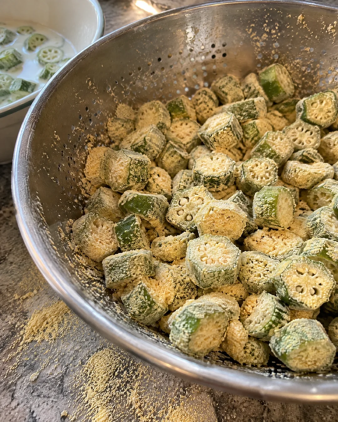 Sliced okra rounds coated in seasoned cornmeal being shaken in a colander so the loose meal falls away