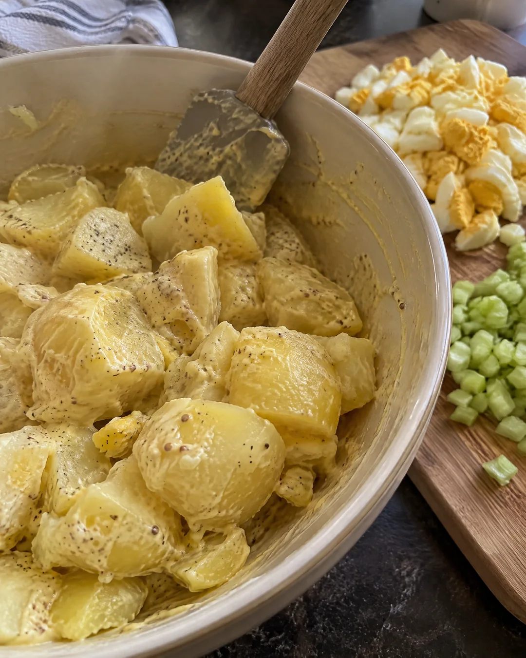 Warm potato chunks being folded into a pale yellow mustard and mayonnaise dressing in a mixing bowl