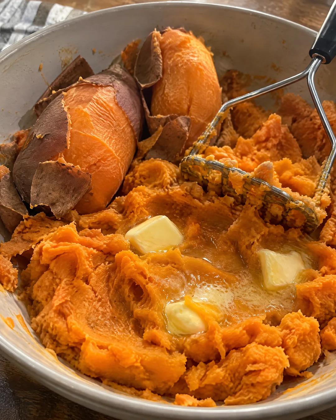 Hot boiled sweet potatoes with their skins slipping off, being mashed with butter in a large bowl until smooth