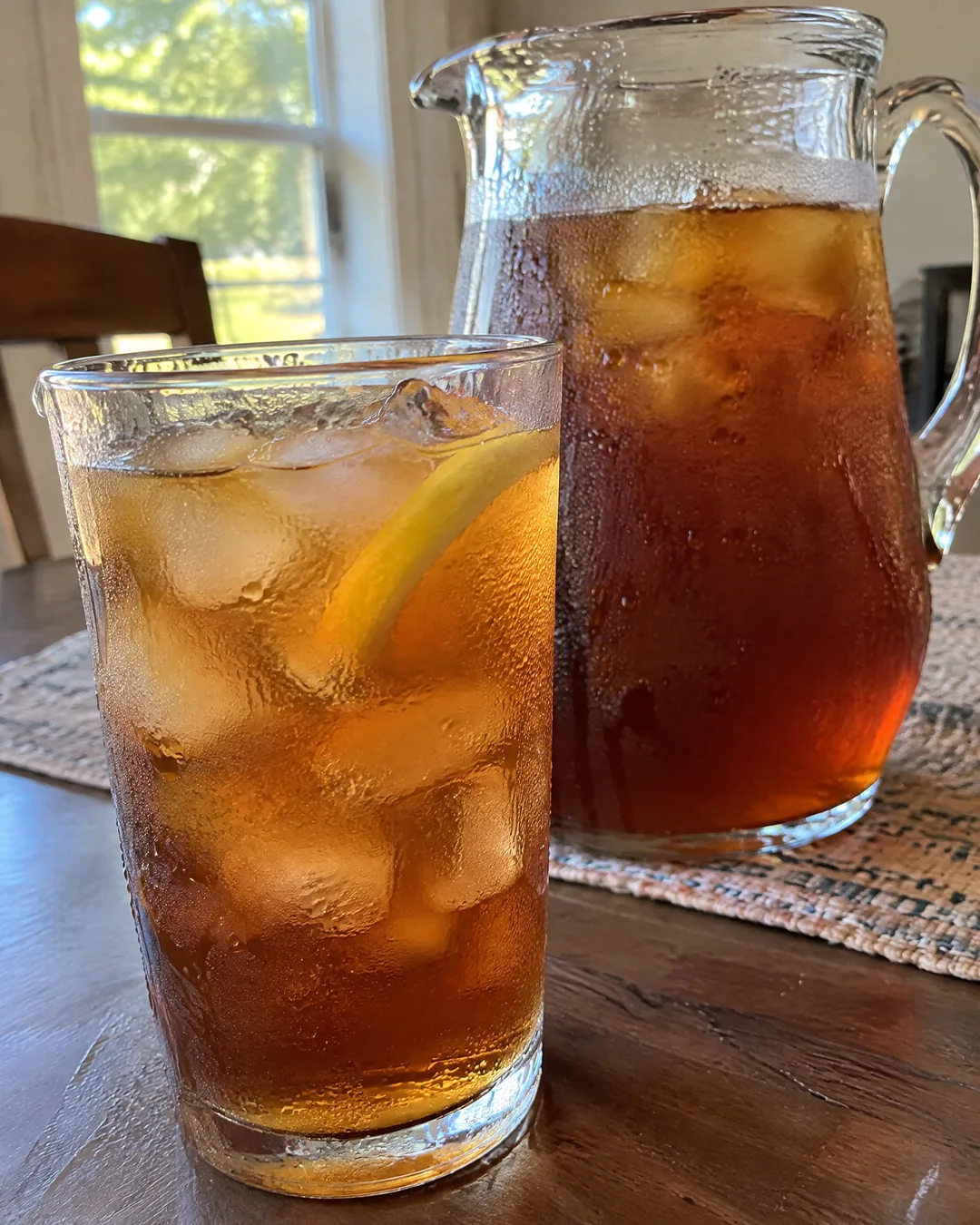 A tall glass of dark amber sweet tea over ice with a lemon wedge, beaded with condensation, beside a full glass pitcher