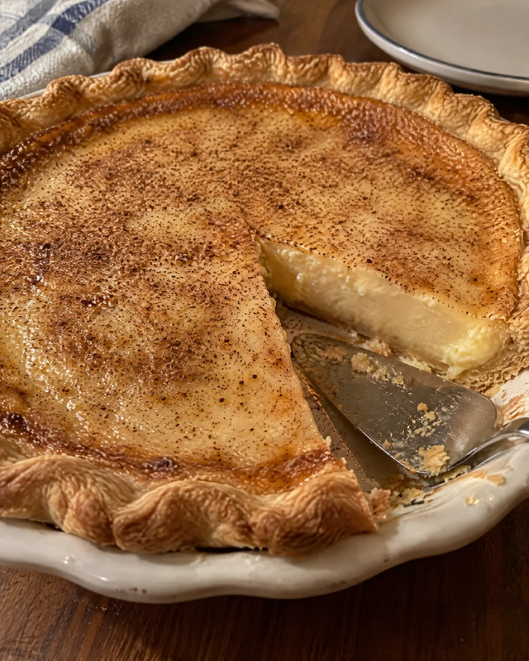 A whole baked sugar cream pie with a golden caramelized skin and nutmeg on top, one slice removed