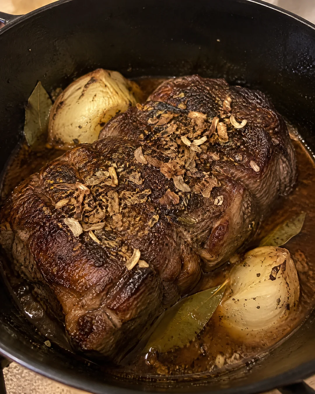 A seared chuck roast in a Dutch oven, dark brown on top, with onion soup seasoning and water around it and onion halves tucked beside