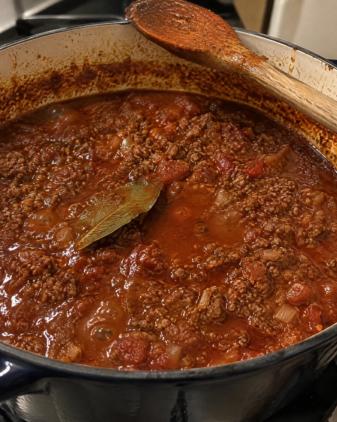 A big pot of thick dark red ground beef meat sauce simmering with a bay leaf and a wooden spoon