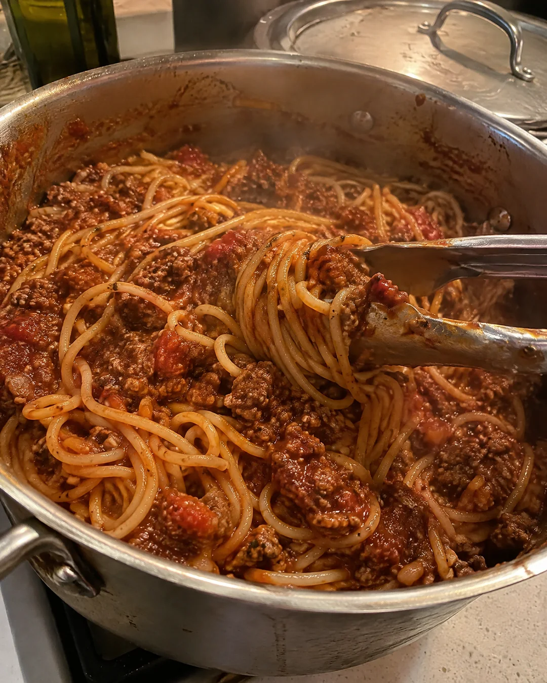 Spaghetti stirred into the pot of meat sauce, every strand coated, with tongs lifting a tangle of noodles