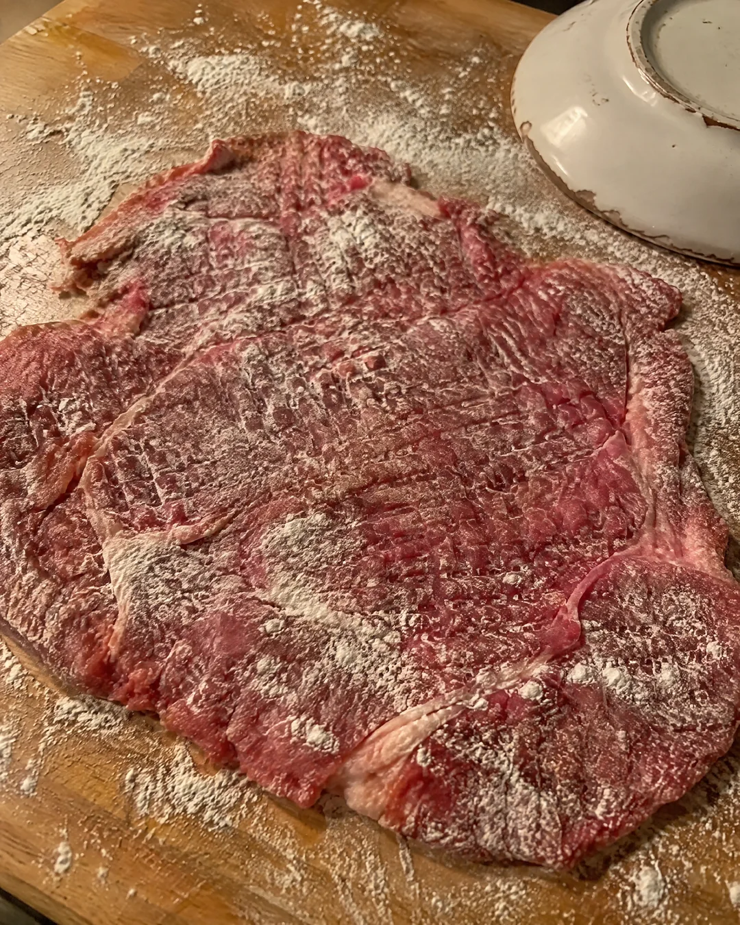 Round steak pounded thin and wide with flour on a wooden board, a heavy saucer resting beside it