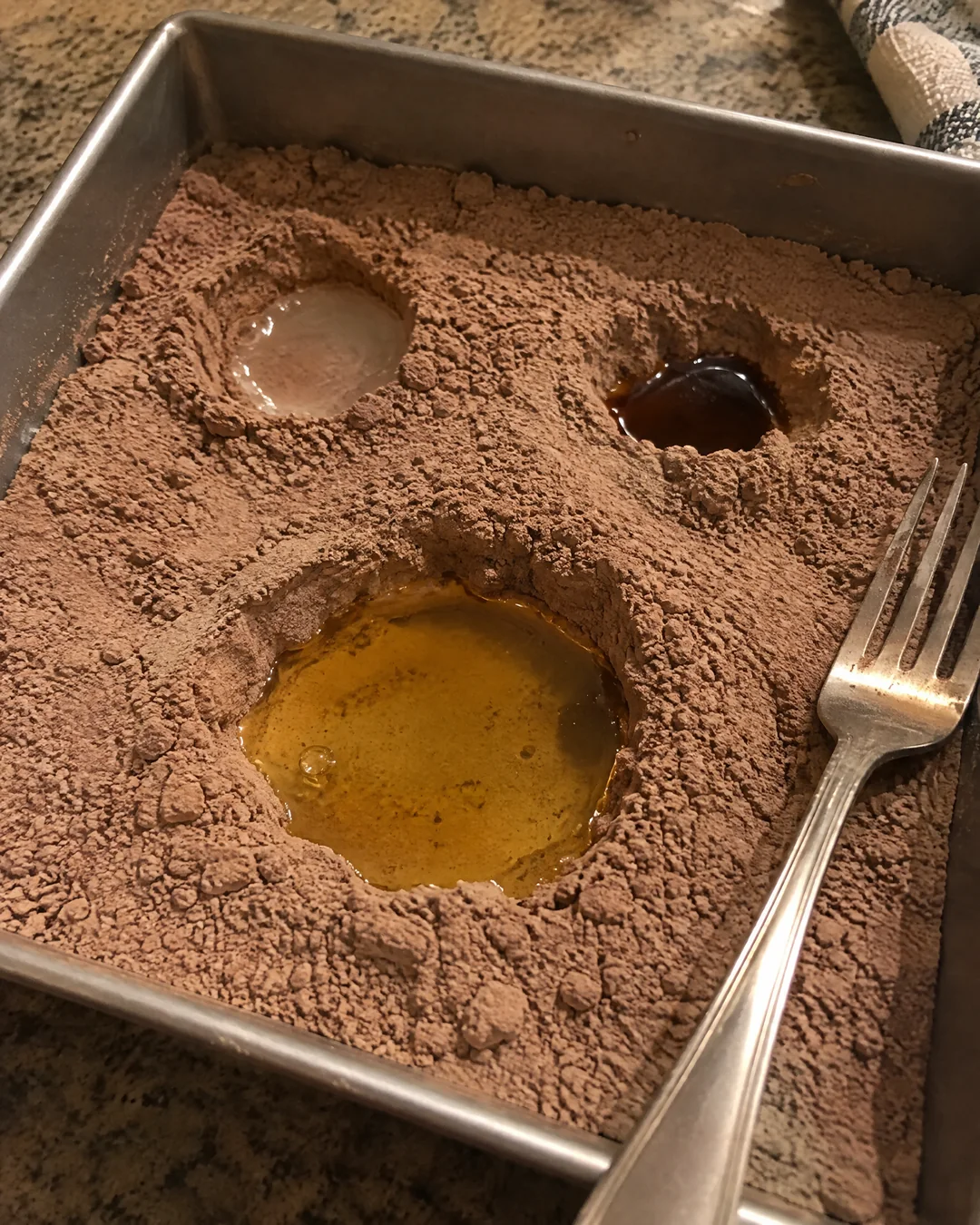 Three holes poked in the dry cocoa mixture in a square pan, filled with vinegar, vanilla and oil before the water goes on