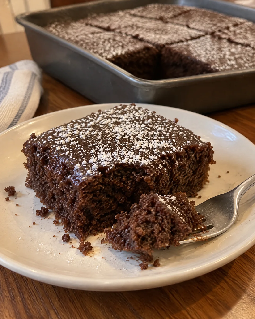 A square of dark moist wacky cake on a plate with a dusting of powdered sugar, the rest of the cake cut in the pan behind