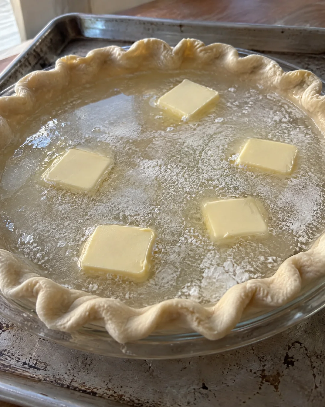 An unbaked deep dish pie crust filled with water, flour and sugar sprinkled over the top and five pats of butter floating, on a baking sheet