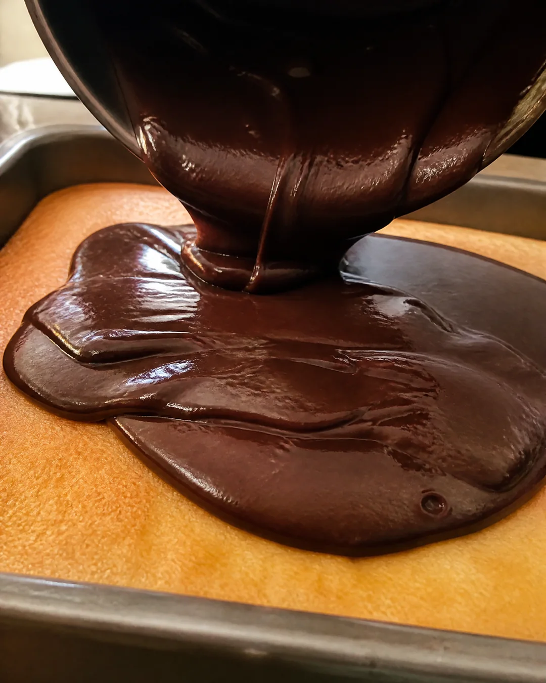 Warm chocolate fudge frosting being poured over a yellow sheet cake in its pan